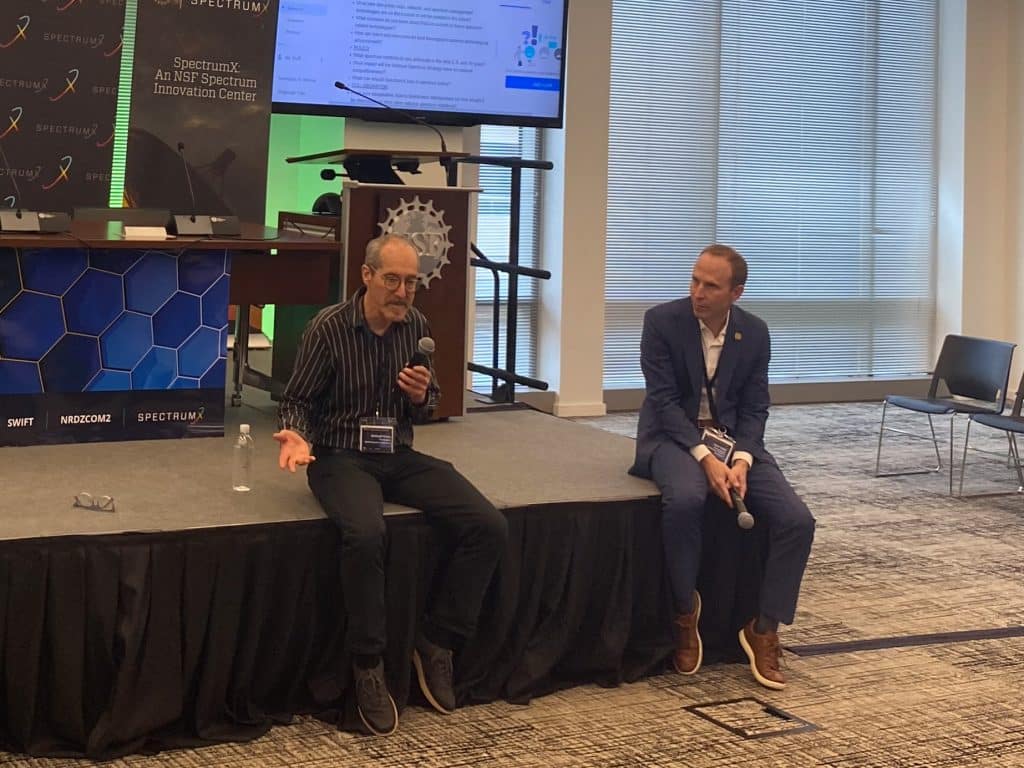 Michael Honig, SpectrumX Research Director and Professor at Northwestern University, sits on the NSF Spectrum Week stage with a microphone in hand discussing research initiatives. His glasses are on the stage next to him. To the right of Michael is Josh Tullis, SpectrumX Managing and Collaboration Director, with his microphone in hand, listening.