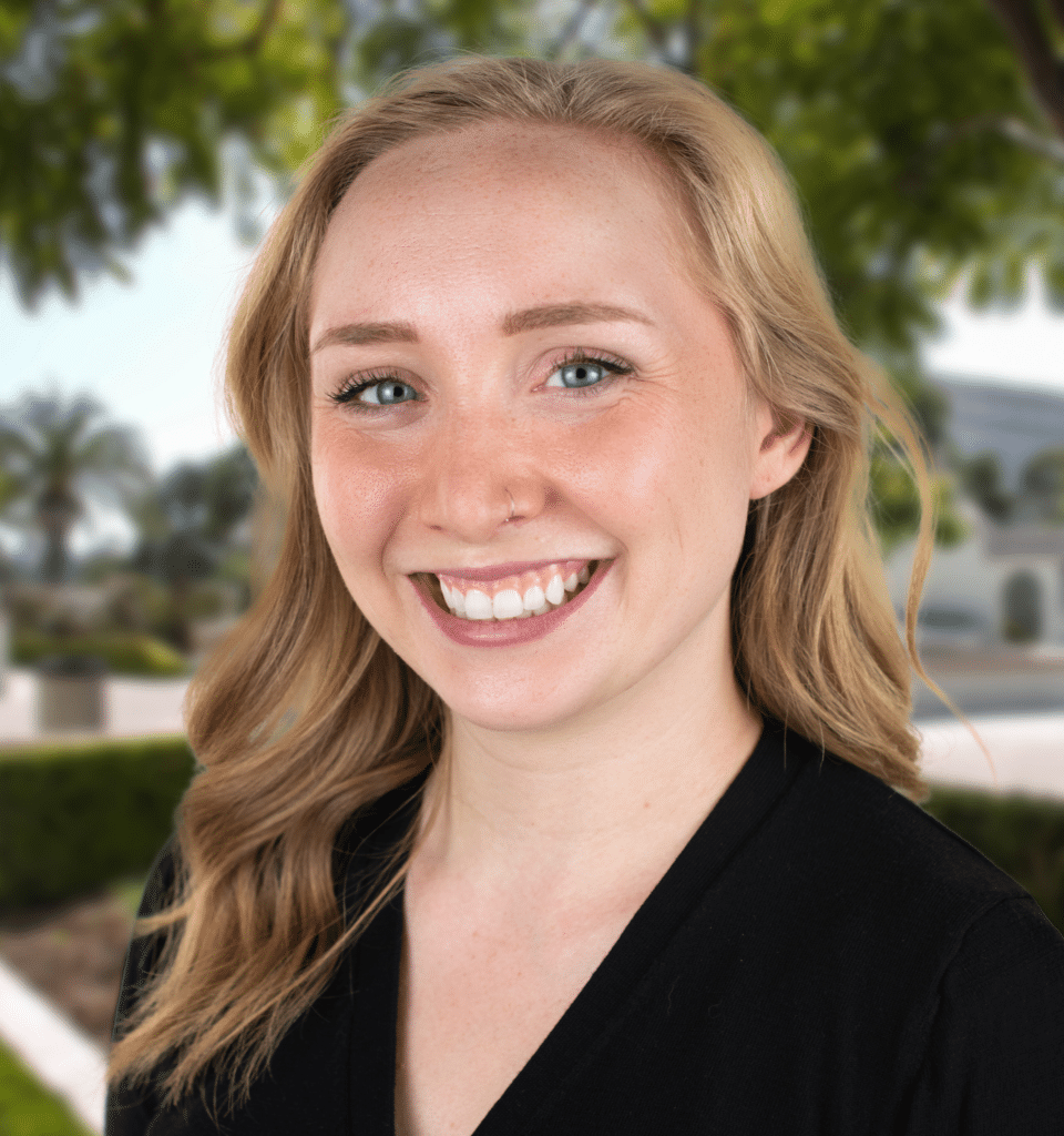 A blonde white woman with blue eyes wearing a black cardigan smiles in front of a background of green leafy trees.