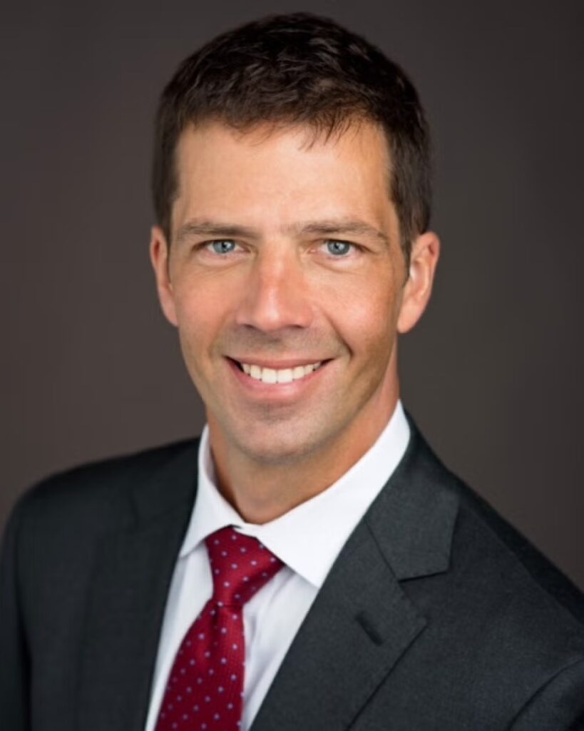 A white man with blue eyes in a black suit, white button up shirt, and red tie smiles in front of a dark brown background.