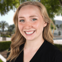 A blonde white woman with blue eyes wearing a black cardigan smiles in front of a background of green leafy trees.